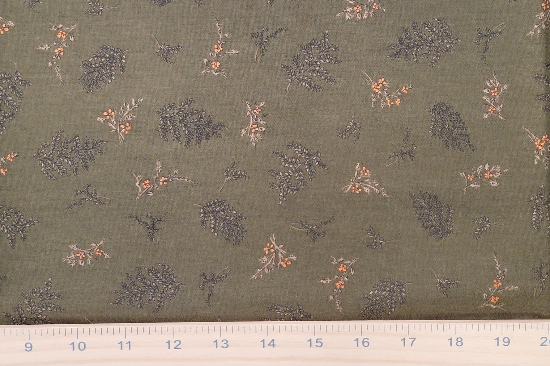A close-up of olive green fabric with a repeating pattern of small gray-green leaves and orange berries, with a white ruler along the bottom edge showing measurements.