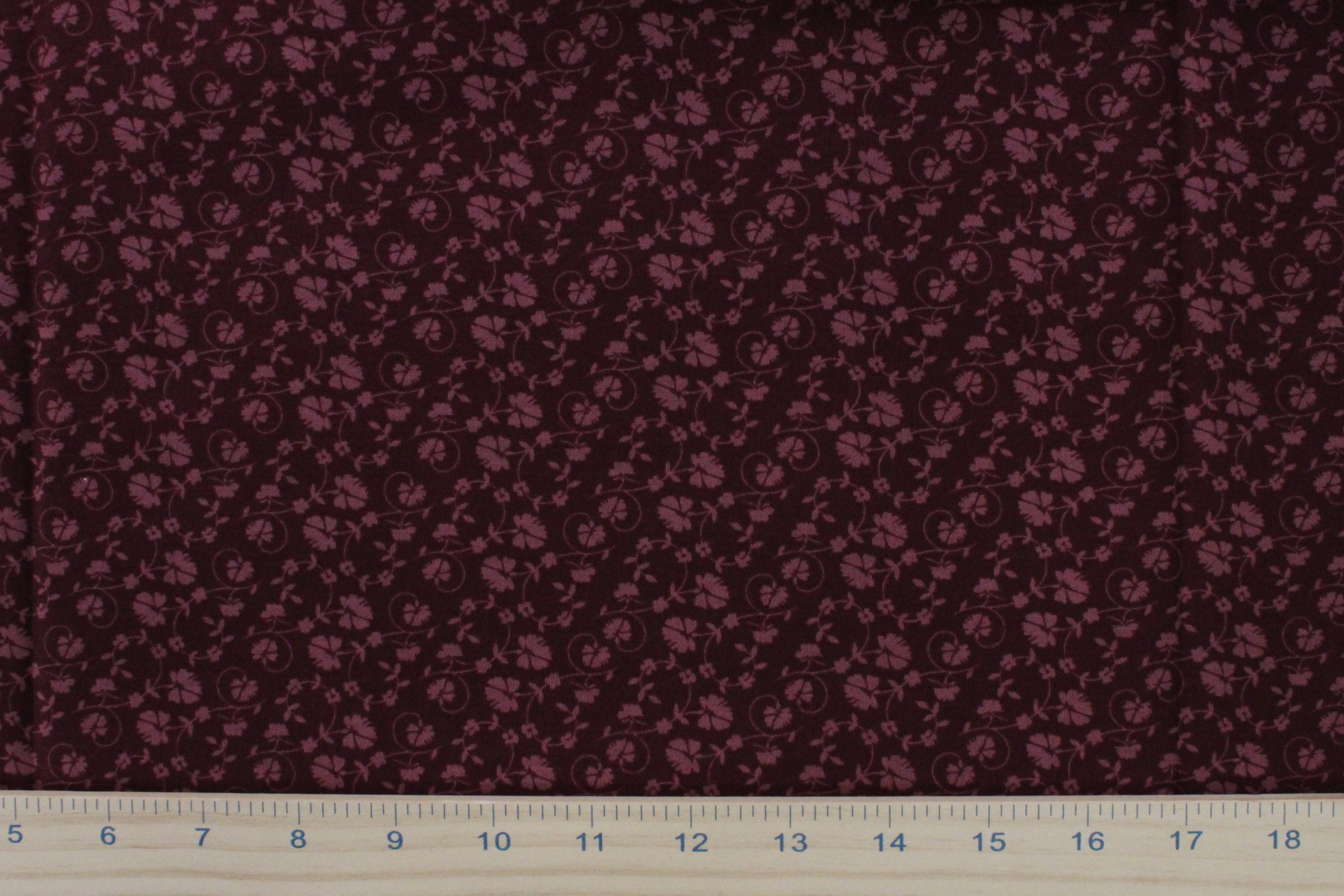 A close-up of dark burgundy fabric with a repeating small floral or paisley-like print in a muted mauve-pink, with a white ruler along the bottom edge for scale.