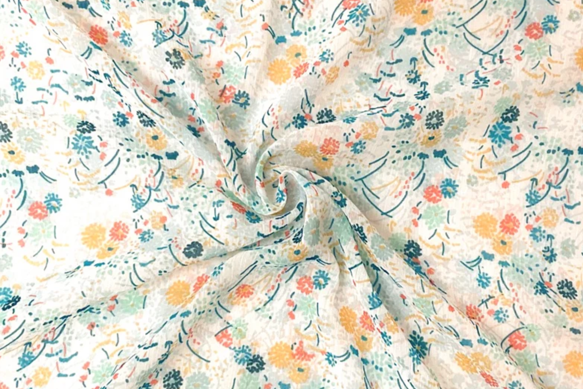 A close-up of a lightweight fabric with a delicate floral print, swirled in the center to show its soft texture and drape. The pattern features small yellow, orange, and teal flowers with green leaves on a warm off-white background.