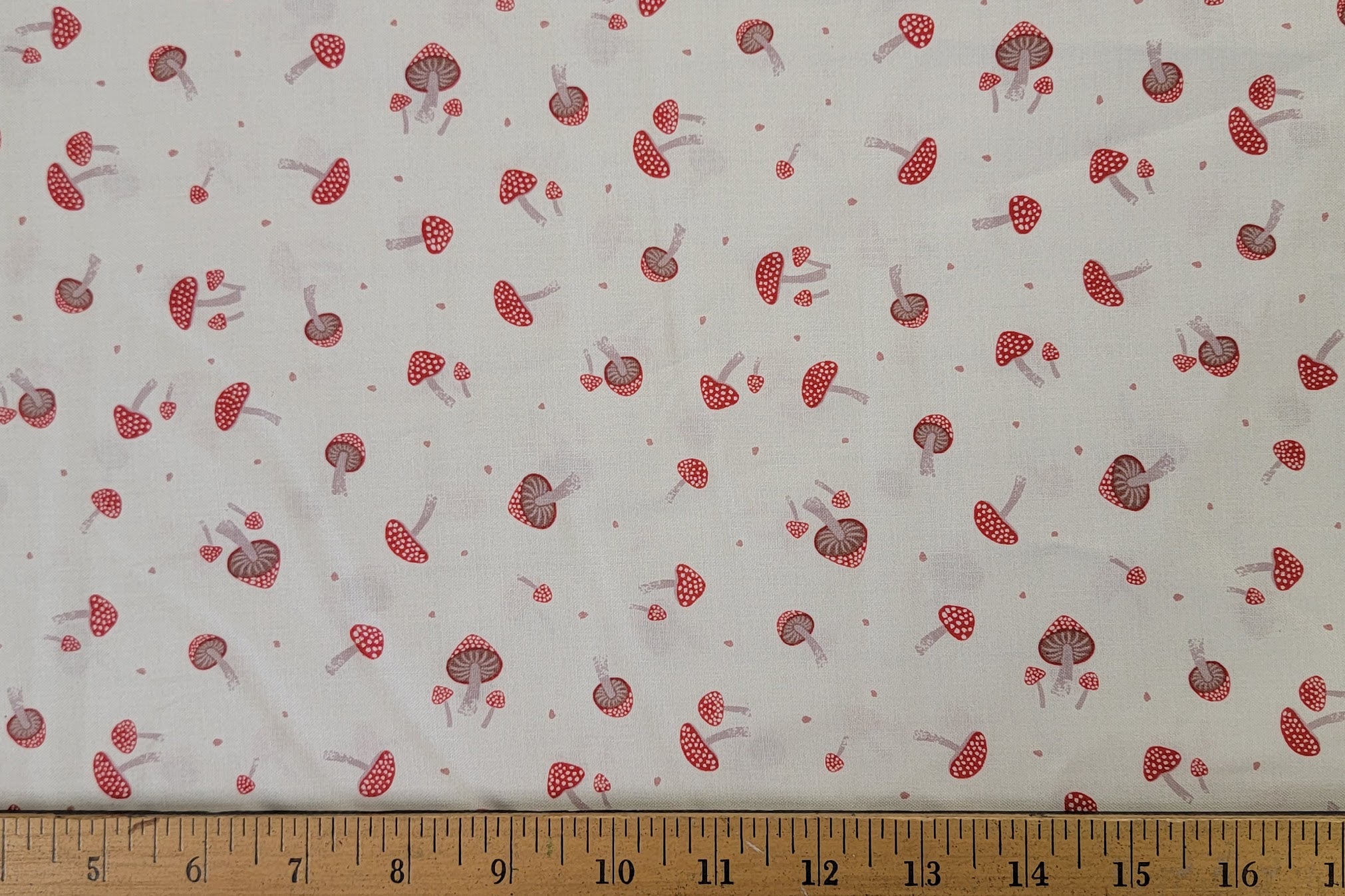 White fabric with a repeating pattern of red and brown mushrooms and small red dots, shown with a wooden ruler along the bottom edge for scale.