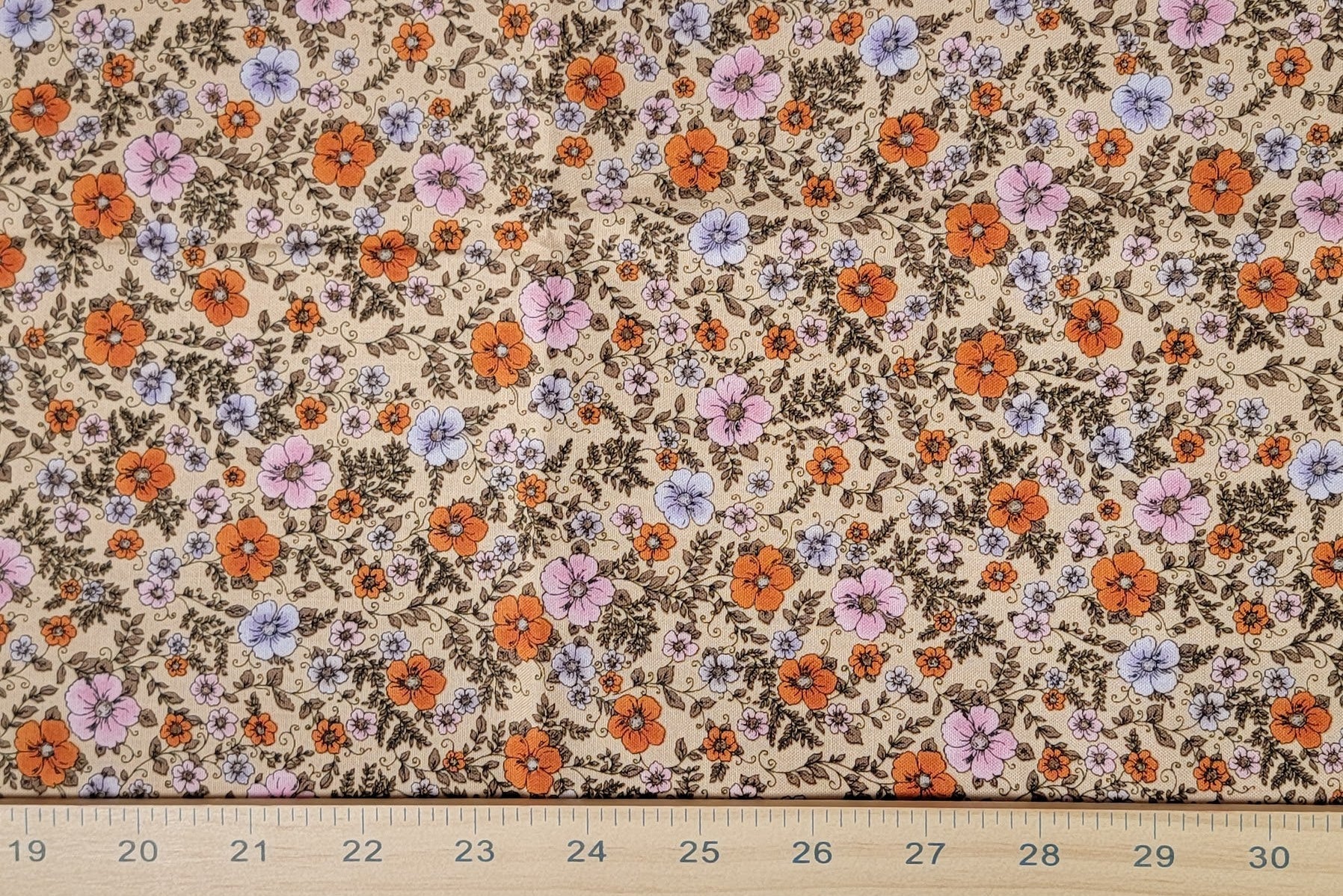 A close-up of a floral fabric pattern with small orange, purple, and pink flowers on a beige background, with a wooden ruler along the bottom edge showing measurements.