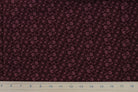A close-up of dark burgundy fabric with a repeating small floral or paisley-like print in a muted mauve-pink, with a white ruler along the bottom edge for scale.