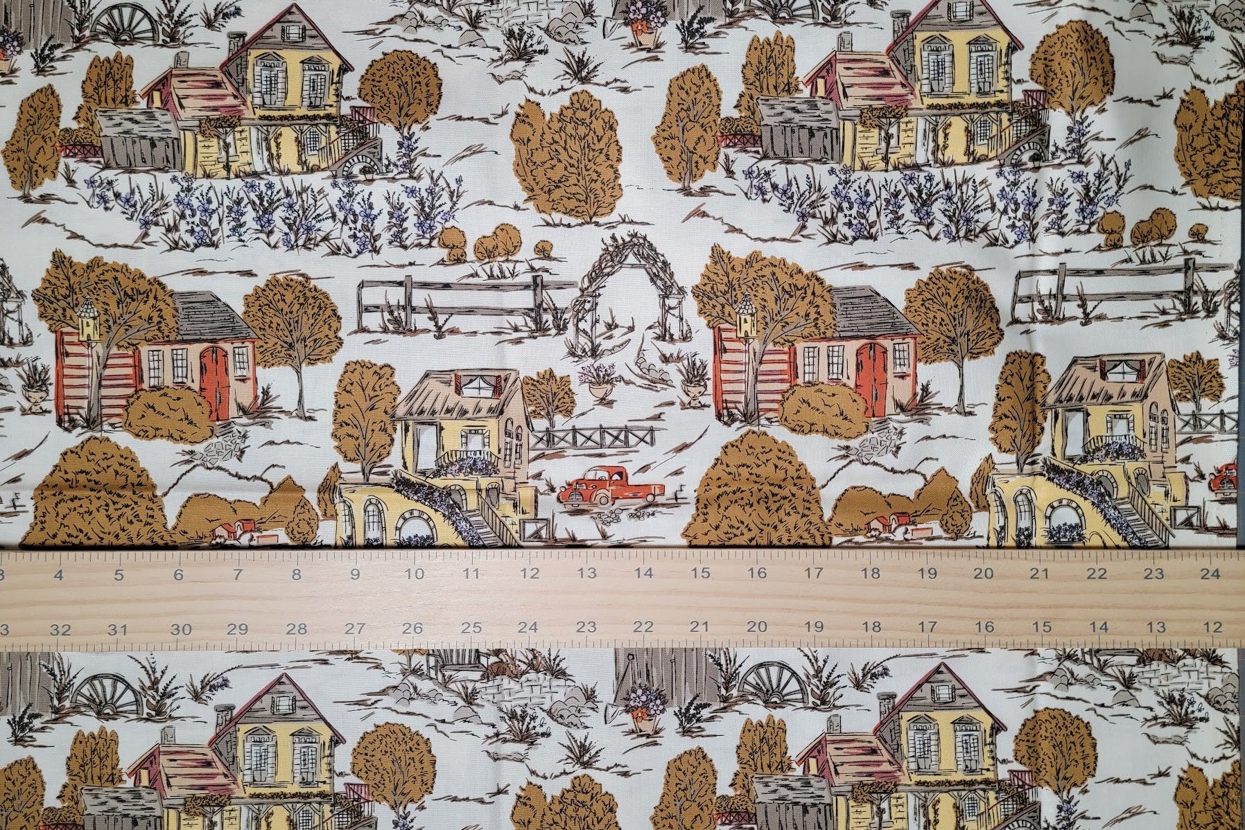 A close-up of a fabric pattern featuring a classic toile design of rural houses, trees, and a red truck on a white background, with a wooden ruler showing measurements across the bottom to indicate scale.