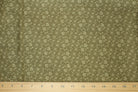 A close-up of a fabric surface with a small repeating floral or paisley-like pattern in muted olive green, with a wooden ruler laid across the bottom for scale.