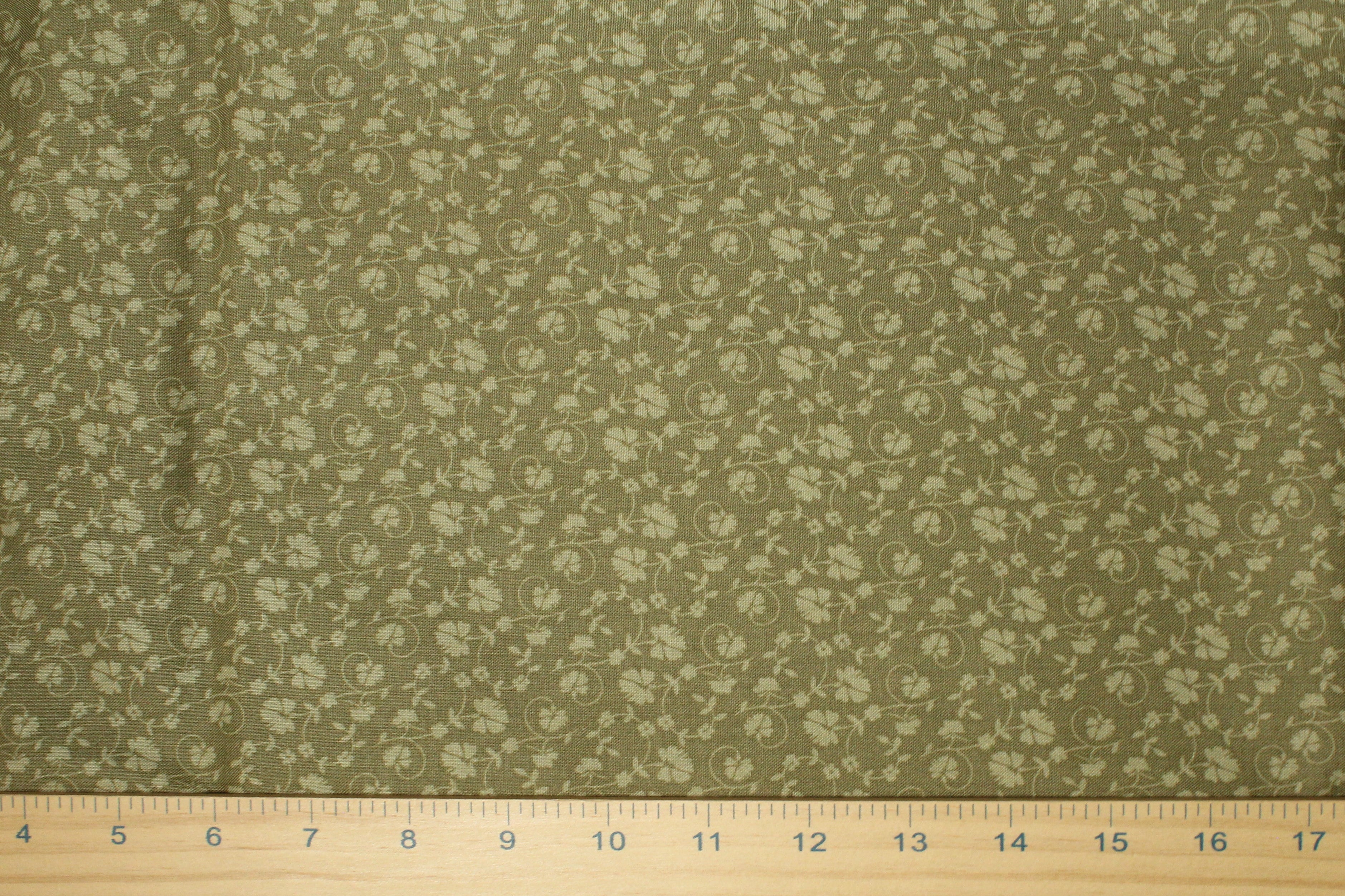 A close-up of a fabric surface with a small repeating floral or paisley-like pattern in muted olive green, with a wooden ruler laid across the bottom for scale.