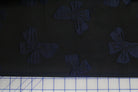 A close-up of a dark fabric with a repeating bow pattern, with a white cutting mat visible along the bottom edge.