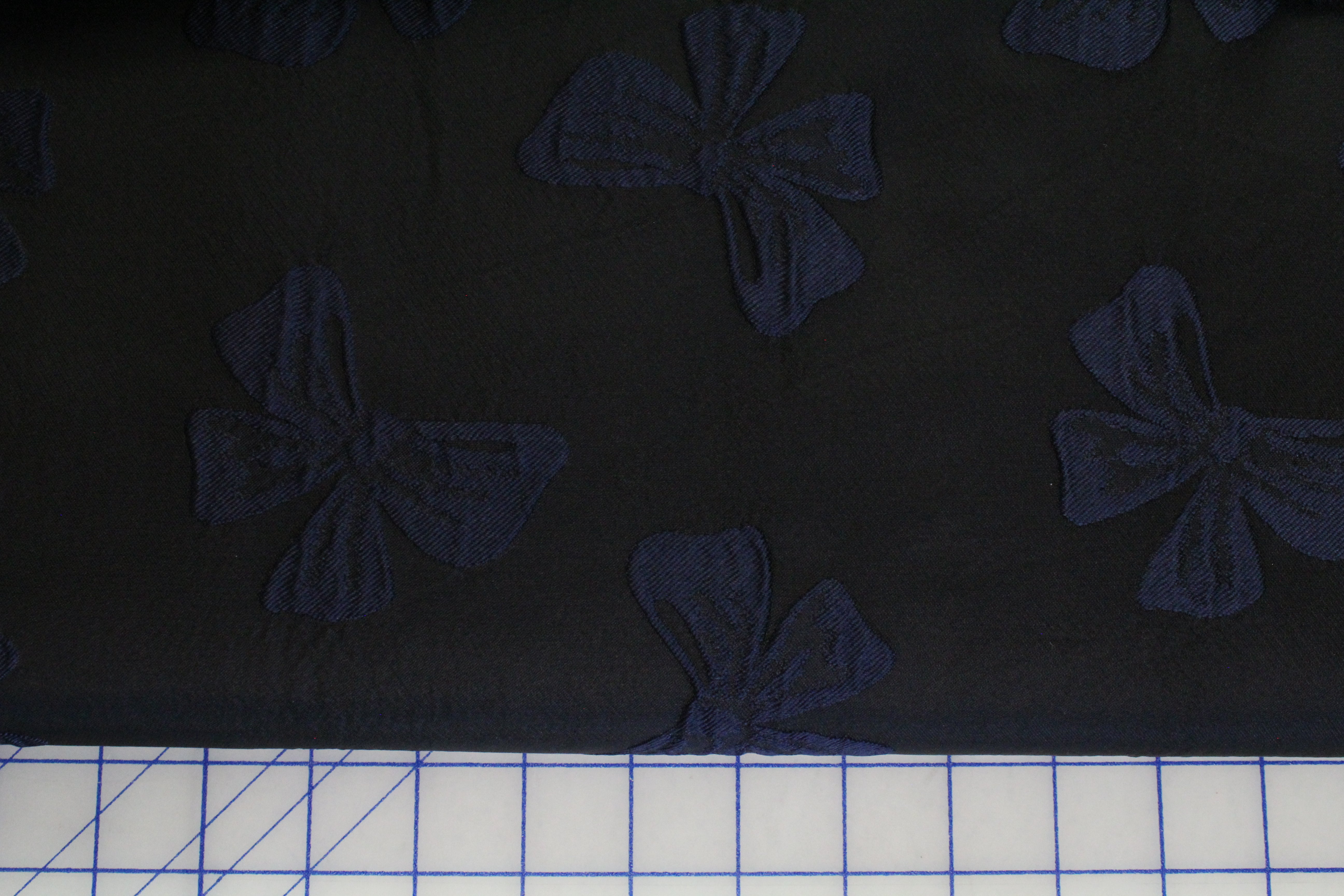 A close-up of a dark fabric with a repeating bow pattern, with a white cutting mat visible along the bottom edge.